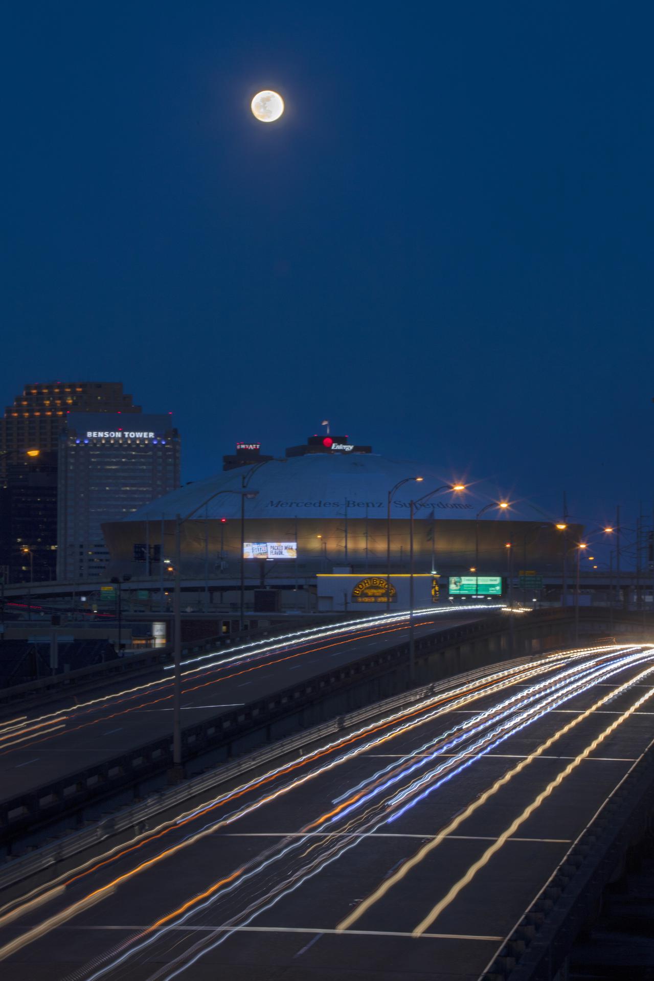 The nearly full moon rises over the city of New Orleans in a time lapse video where the headlights of cars are streaks on the road.