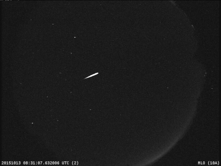 A bright meteor streaks across a dark sky in front of a crescent Moon. 