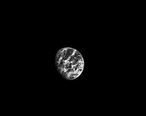 Three-fourths of Earth is seen in black and white, suspended in the blackness of space. Earth only takes up a small portion of the center of the image, and wispy clouds are visible above its surface.