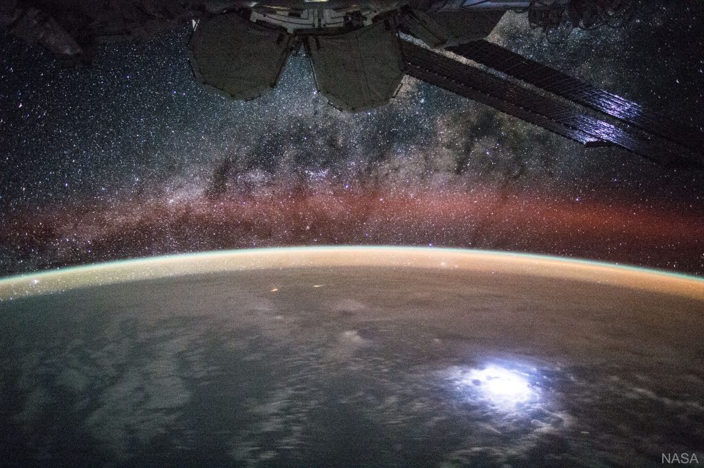 The bottom half shows a dark, cloudy Earth from above. The middle of the image, outlining the Earth is a glowing line, that goes from orange, to teal green. Above the Earth, there is a dark starry sky. There is a red foggy line – airglow. Peaking out from the top is the International Space Station.