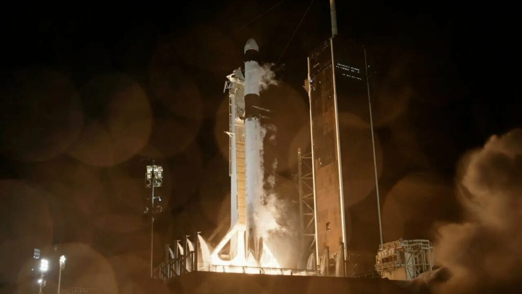 At night, a large, white rocket stands straight up in the middle of the image. On either side are tall towers. The bottom of the rocket is bright, as if it's lighting up, and golden smoke starts to fill the bottom of the image.