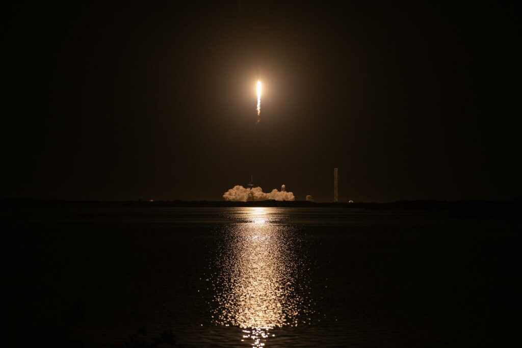 Set against a black sky, a rocket rises above a launch pad with a bright yellow glow and trail of white smoke directly beneath it. Plumes of white smoke billow around the launch pad on the ground. In the foreground, the light from the rocket engine is reflected in dark waters.