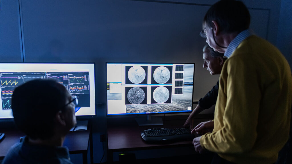 Three men stand in a darkened room, in front of two computer monitors with images of data on the screens. 