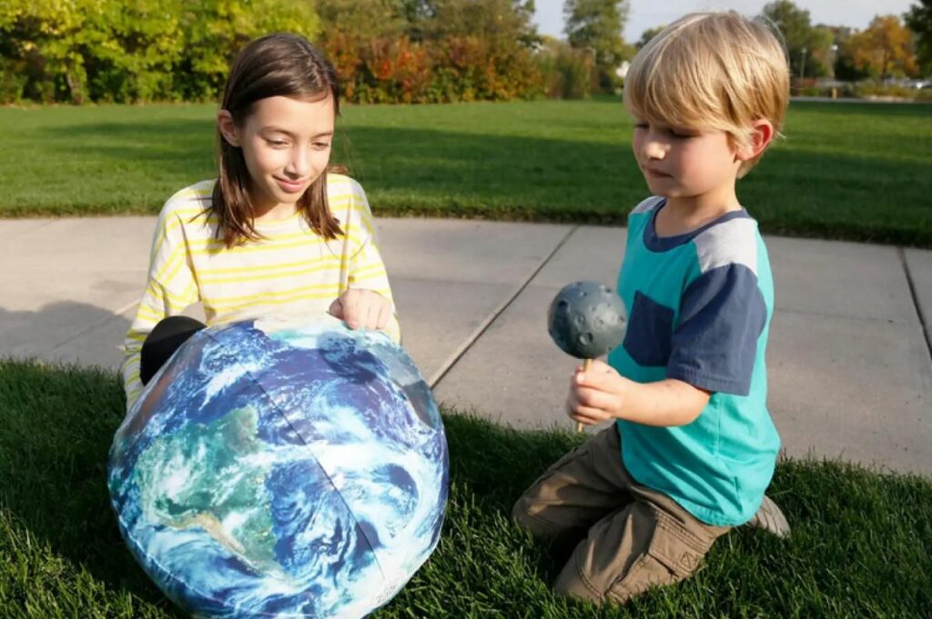 Two children demonstrating an eclipse with an Earth-printed beach ball and a Moon-shaped ball.