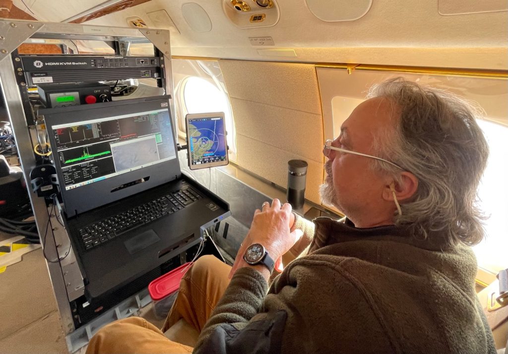 On the aircraft a scientists sits in gront of a boxy instrument, looking at the screen readout.