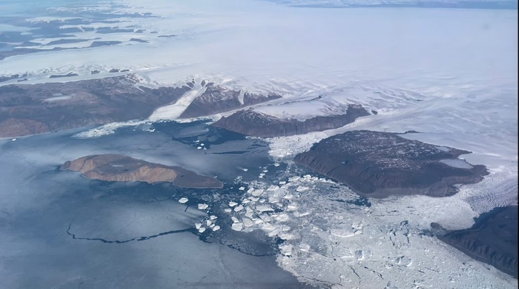 A view from the plane of Grenland's white glaciers on land in the top and top right of the image. Brown rock of the cliffs and shoreline are a buffer between the land ice and the gray-blue sea ice on the ocean. Broken chunks of icebergs are on the right edge of the image, and a brown rocking island is in the bay.