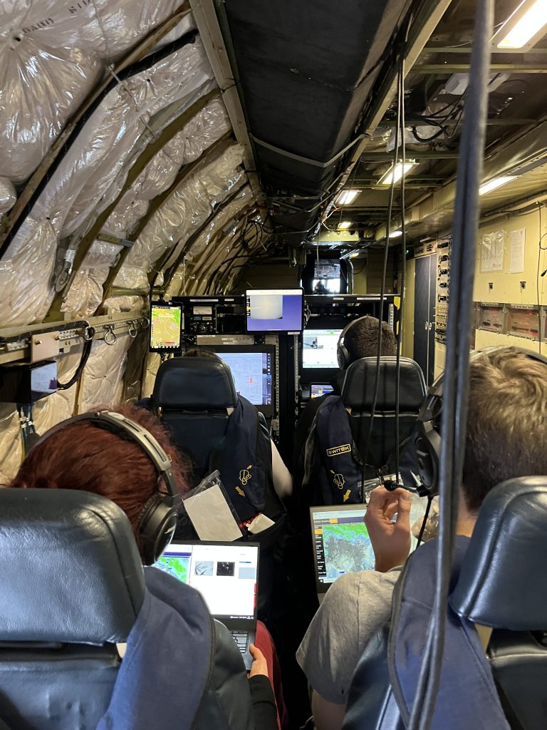 Four scientists strapped into their seats, focusing on the scientific data on the monitors and laptops in front of them. The photo is taken from behind them. the scientists are sitting in what appears to be the inside of a passenger plane that has been gutted and filled with scientific instruments.