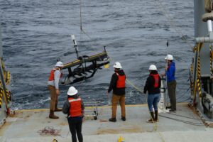 A group of researchers, equipped with hard hats and life vests, pull at ropes to assist the S-MODE waveglider lower into the ocean.