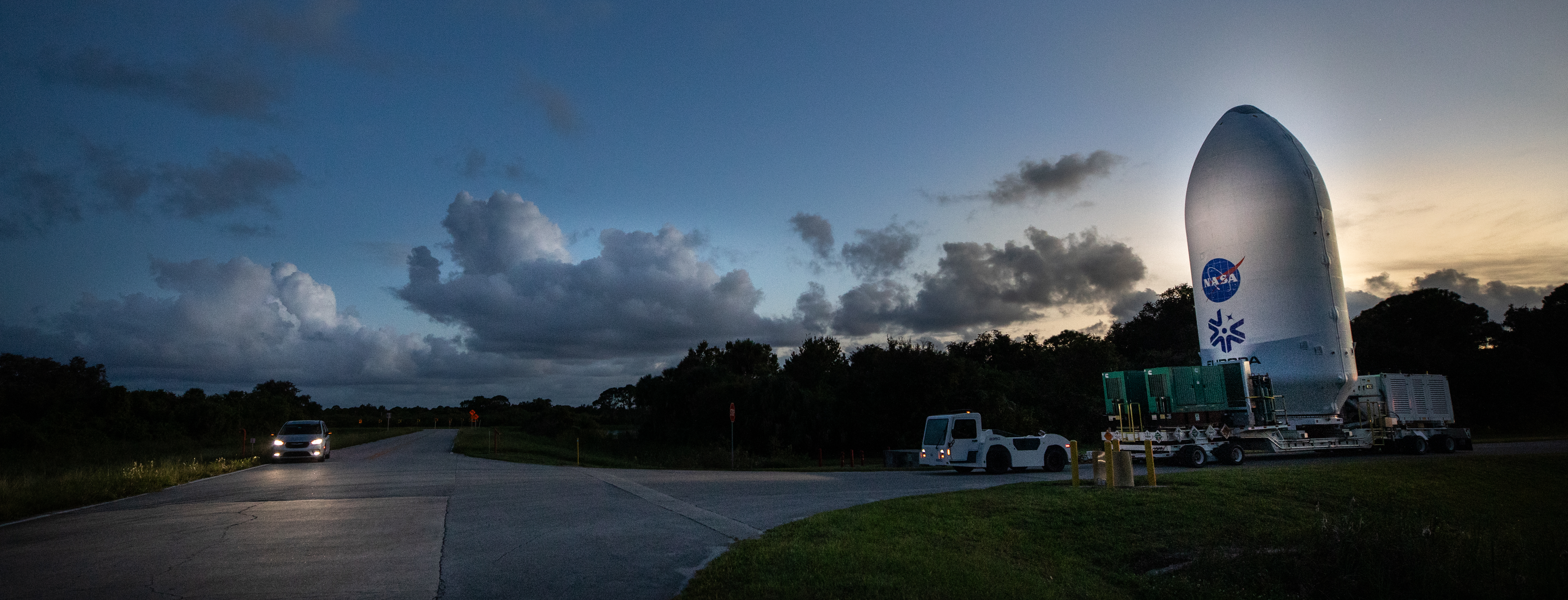 A transport truck pulls a SpaceX payload fairing away from the processing facility at dusk. The fairing has the NASA logo and Europa Clipper emblem printed on the front. The sun has set leaving a colorful glow on the horizon. 