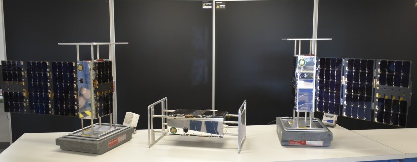 Three small silver and black satellites are displayed in a row on a table. The two satellites on the left and right stand vertically and each have one solar array extended. The satellite in the middle is displayed horizontally and has its solar arrays folded up.