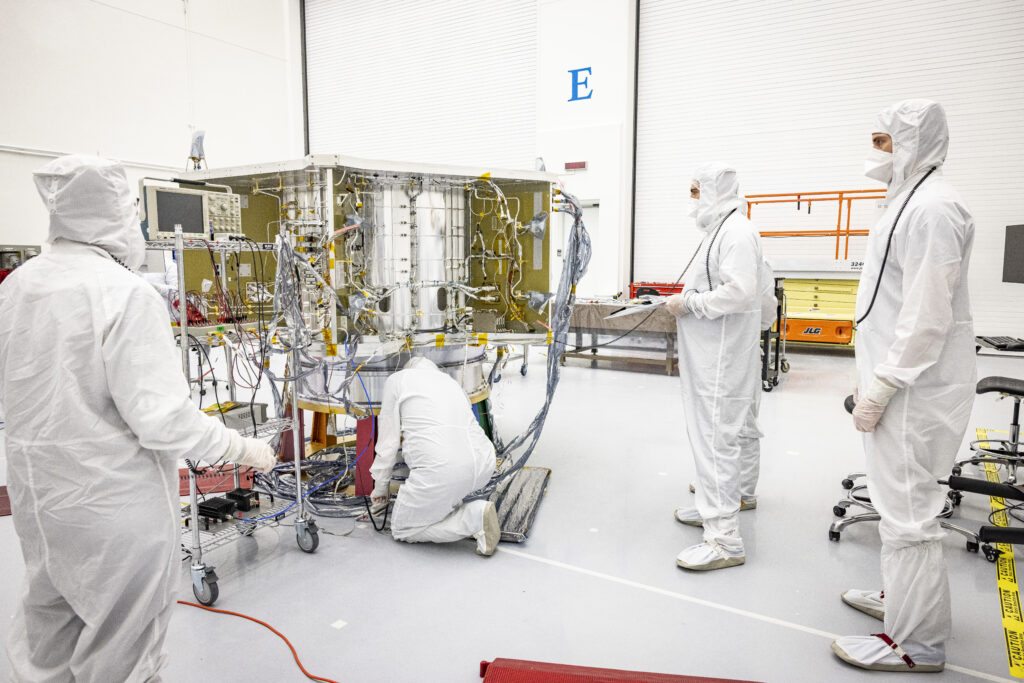 Four people in white suits and masks are in a white clean room, three are standing and looking at a piece of equipment covered in various wiring. The fourth person is kneeling and working next to the equipment.