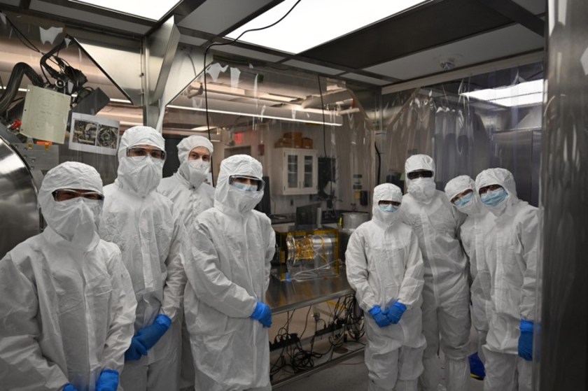 Eight people in white clean-room suits and masks stand around the gold, black, and silver SWAP instrument that will be installed on the IMAP spacecraft.