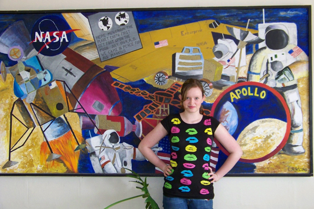 Erica Kriner as a child, posing in front of an abstract art mural that consists of NASA, Apollo, and other space-related items.