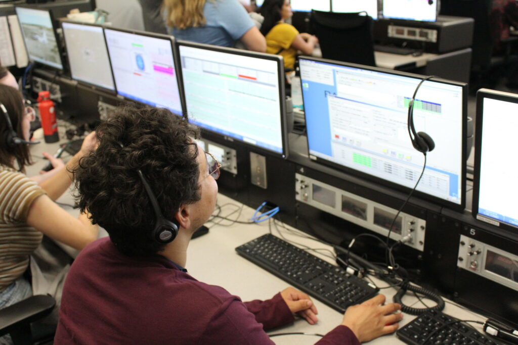 A group of NASA Community College Aerospace Scholars sit in the control room at NASA Armstrong Flight Research Center. Each of the scholars are at computer working a live simulation of a mission control. The diverse group of interns wear business casual attire. Credit: NASA