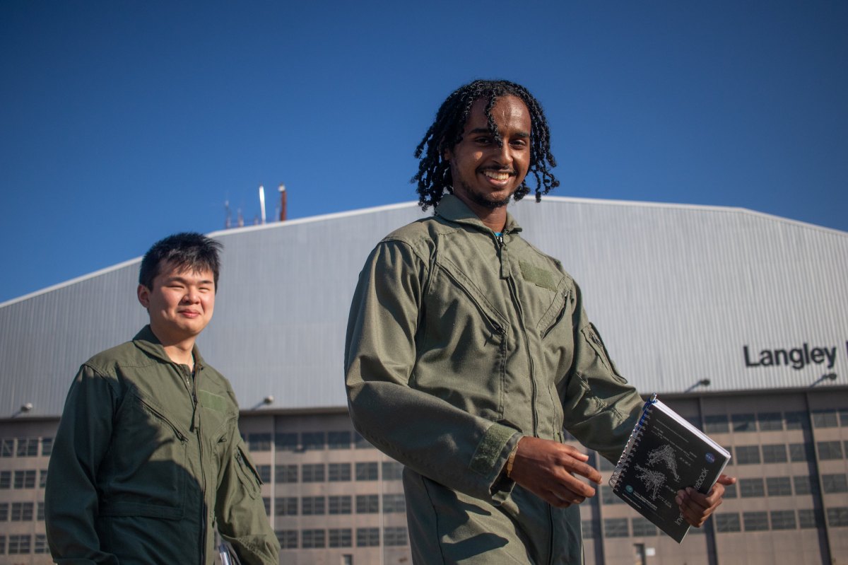 From left to right, NASA SARP Interns Dennie Truong and Nathan Tesfayi exit the NASA Langley Hangar to board the NASA B200 aircraft for their first atmospheric test flights of the summer. Both interns wear brown flight suits and smile with the large hangar and bright blue sky in the background. Credit: NASA/Angelique Herring