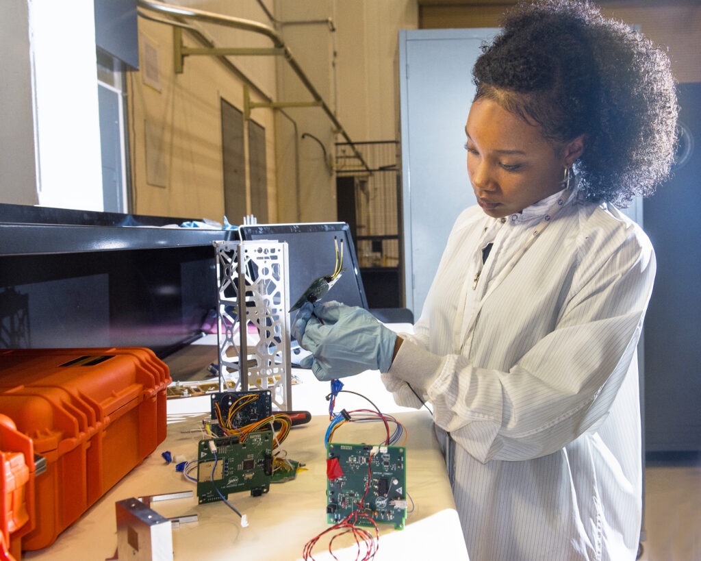 Former NASA intern Naia Butler-Craig works to assemble a CubeSat in her laboratory. Wearing clean room attire she pays close attention to different cabling while assembling the small cube shaped satellite. Credit: NASA/ Bridget Caswell