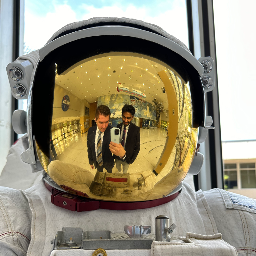 Two NASA interns pose together for a selfie by taking an image of the reflective visor of an extravehicular activity spacesuit. The rounded visor creates a fisheye effect on duo wearing business professional attire. Credit: Matt McDonnell/NASA