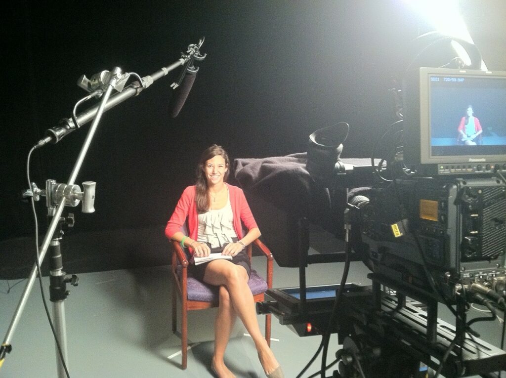 Alt Text: Former NASA intern and mentor Erin Kisliuk sits in an interviewing chair inside the film studio at NASA Headquarters. Erin wears business casual attire and smiles at the camera. In the foreground a large video camera records her. To the left of the foreground, a boom microphone hangs overhead. Credit: Erin Kisliuk