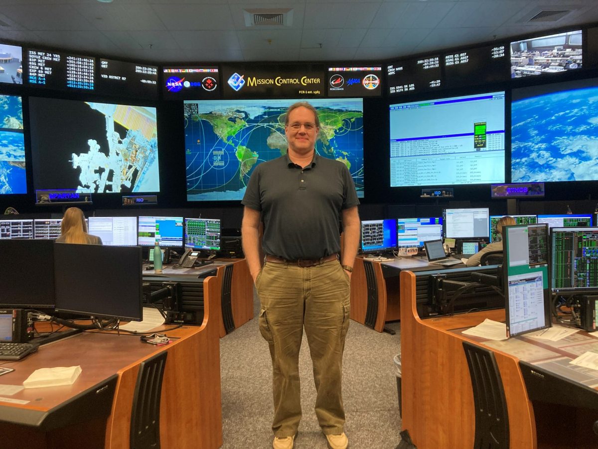 Alt Text: Mike Fogg, a tall white man with glasses and brown hair, smiles at the camera in a gray polo and khaki pants with his hands in his pockets. Behind Mike is a set of screens showing ISS Mission Control. Photo Credit: Nancy Ariza.