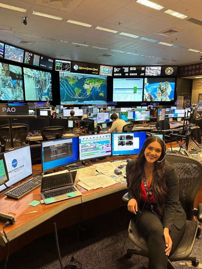 Alt Text: Dominique Crespo, an intern at NASA's Johnson Space Center, wears a headset and smiles while sitting at a console in the Mission Control Center. Behind her numerous computer monitors take up a desk and the background is dominated by massive screens displaying data and information about the International Space Station. 