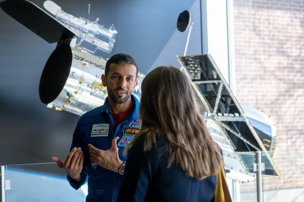 Alt Text: NASA intern Julia Tilton interviews UAE astronaut Sultan al-Neyadi. al-Neyadi wears the iconic blue jumpsuit of an astronaut and gestures with his hands. Tilton is facing away from the camera and speaks with al-Neyadi in business professional attire. In the background a large model of the Hubble Space Telescope is on display. Credit: NASA