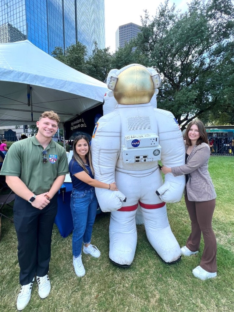 Alt text: Berlie poses with two of her fellow interns tabling for Houston Energy Day. Berlie wears white sneakers, brown flared leggings, and a striped blazer and stands to the right of the photo, holding the arm of a tall inflatable astronaut in a white and red space suit and a golden helmet between the three.