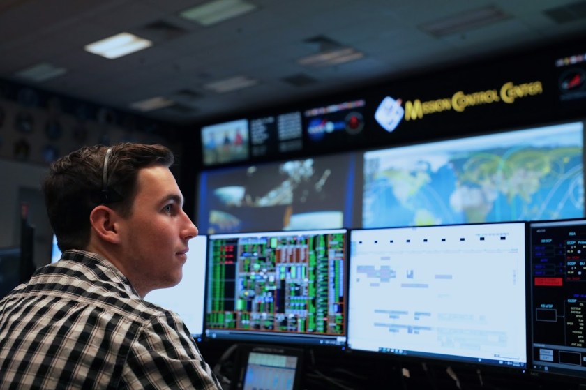 A NASA intern in business casual attire sits in the Mission Control Center at NASA's Johnson Space Center. Looking forward at the console of computer monitors with data. In the background the large screens of Mission Control dominate. Credit: NASA