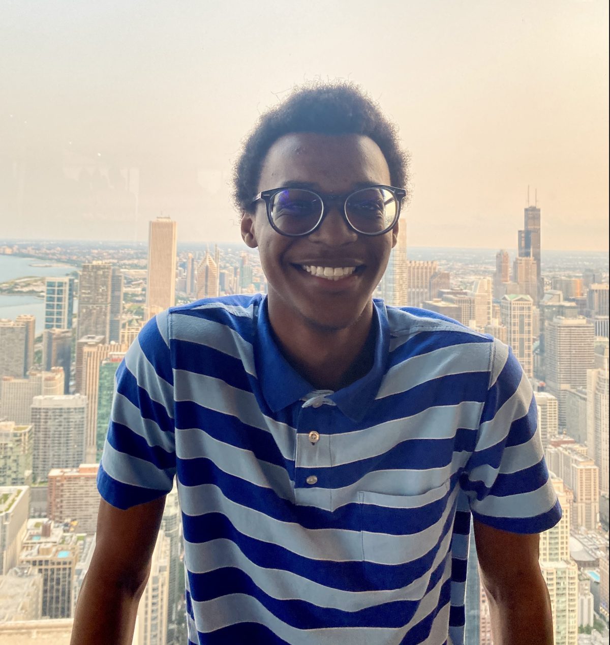 Image Description: Dominating the center of the image, a Black man with gray glasses and a blue striped polo stands in front of a window. Behind him is the New York City skyline and faint orange clouds. Image Credit: Stephan Bellamy.