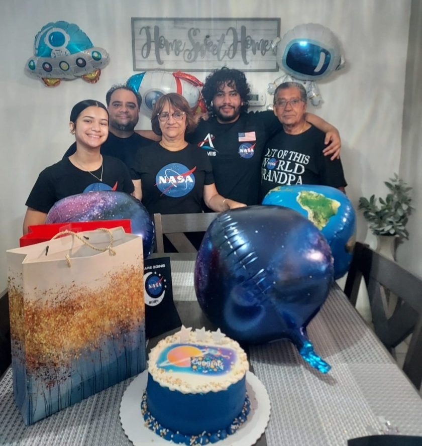 Alt text: Lopez poses with four of his family members celebrating his acceptance as a Summer 2024 NASA intern. The whole family is wearing black NASA t-shirts. In the foreground of the image is a cake with planets on it, a gift bag, and blue balloons. Credit: Johnpaul Lopez