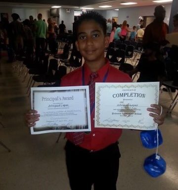 Alt text: In this photo taken in 2014, Lopez poses with two awards. Lopez is in the center of the image wearing a red button-up shirt with a blue lanyard, red tie, and black pants. Credit: Johnpaul Lopez