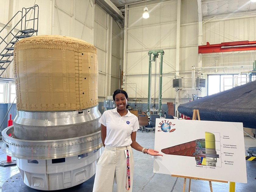 Alt Text: NASA intern Caitlyn McClanahan poses with NASA's LOFTID (Low-Earth Orbit Flight Test of an Inflatable Decelerator) within a large hangar at NASA's Langley Research Center. She smiles and wears business casual attire. Credit: Caitlyn McClanahan