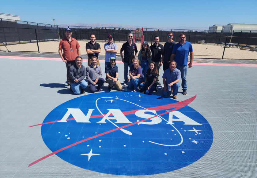 Alt Text: Nesto Cano, alongside a diverse team of colleagues, pose atop a red, white, and blue NASA meatball logo.