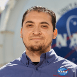 NASA Intern Nestor Cano smiles in front of a large metallic structure emblazoned with the iconic red, white, and blue NASA "meatball" logo. Wearing a button-down shirt with the NASA logo embroidered he stands outside on a sunny day. Credit: NASA