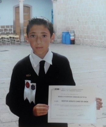 Alt Text: Nestor Cano as a young student. He poses alongside a certificate of achievement and two ribbons he received. He wears a school uniform with a dark tie and matching sweater. 