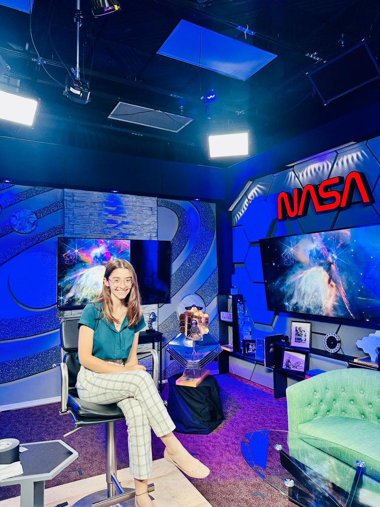 Alt Text: Nora Lowe smiles at the camera in a teal NASA polo and patterned pants. She is seated in a chair positioned to the left of the room, with a television behind her and another on the right, showcasing images of galaxies. Above the right screen, the iconic red NASA worm logo stands out. The room is bathed in deep blue lighting from the ceiling, while a vibrant purple glow radiates from the floor, creating a cosmic ambiance. Image Credit: Nora Lowe