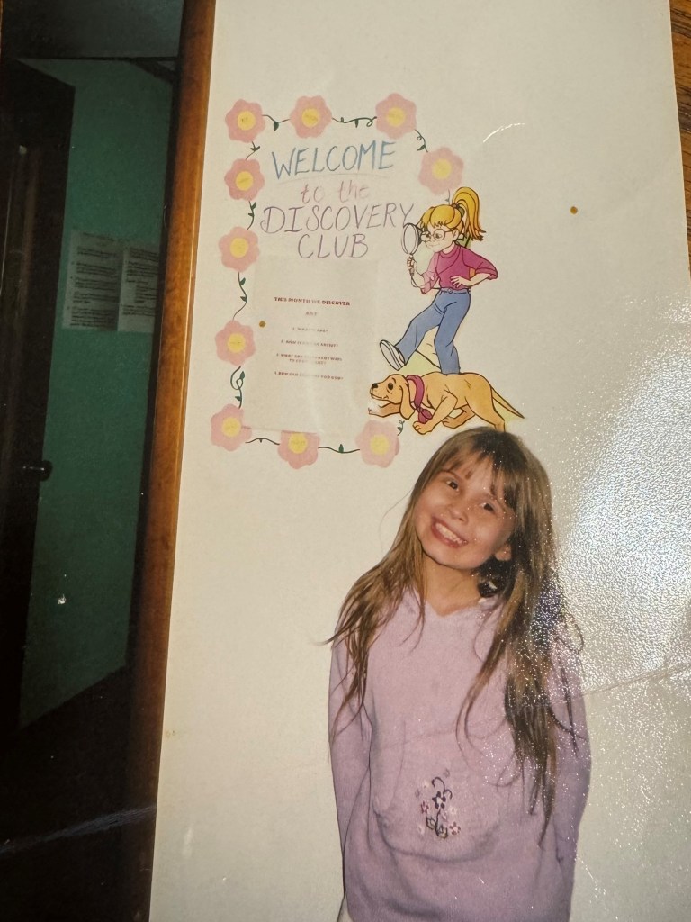 Alt Text: A young Tara Roanhorse smiles infront of a poster on the wall reading “Welcome to the Discovery Club”. Tara wears a light blue sweatshirt with flower embroidery and smiles. Credit: Tara Roanhorse