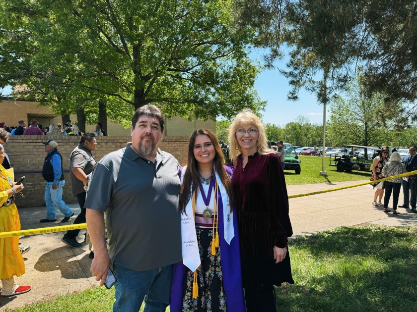 Alt Text: NASA Intern Tara Roanhorse smiles standing with her mother and father after receiving her Associates of Arts degree. She wears a traditional tribal dress and necklace along with her graduation robe and stole as well as multiple honors chords around her neck. Credit: Tara Roanhorse