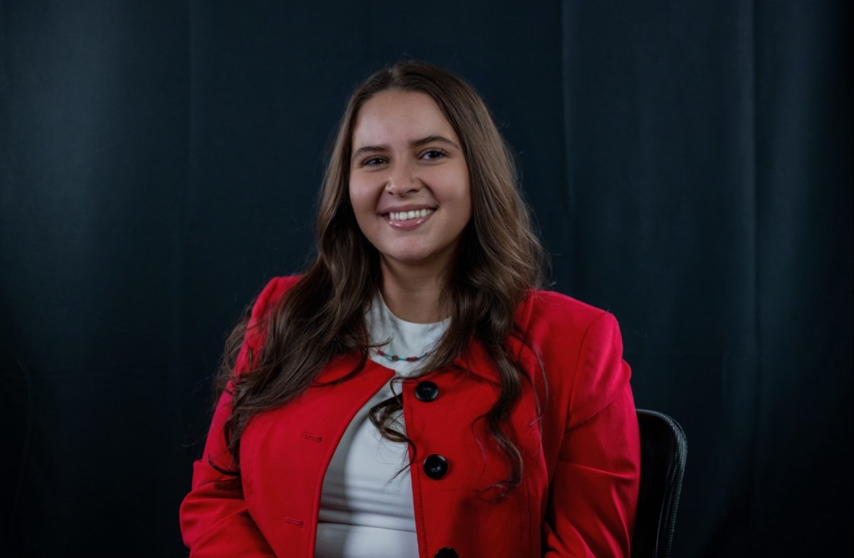 Alt Text: Tara Roanhorse, an indigenous woman with tan skin and brown hair, poses for a portrait in a red jacket, white shirt, and bead necklace. Credit: Tara Roanhorse