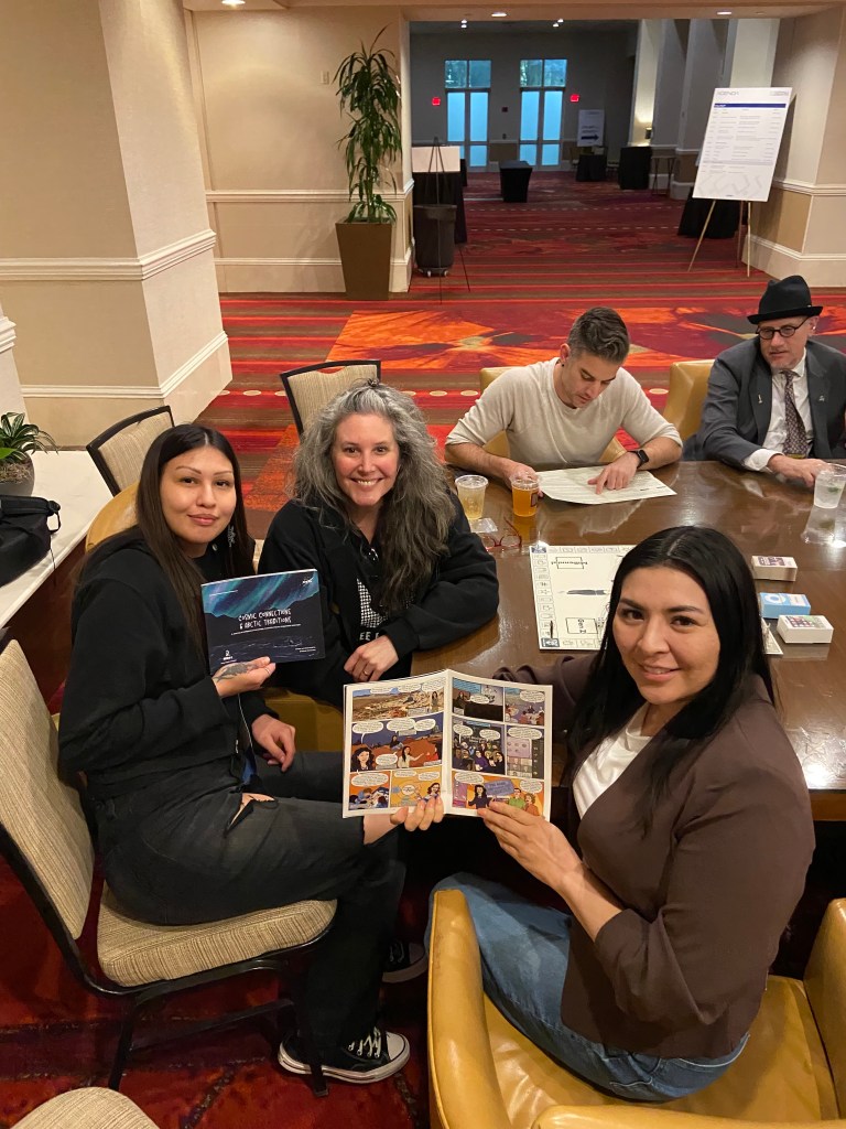 Alt Text: Caitlin Bordeaux and her team sit together at a large table admiring the comic book pages they collaborated on. 