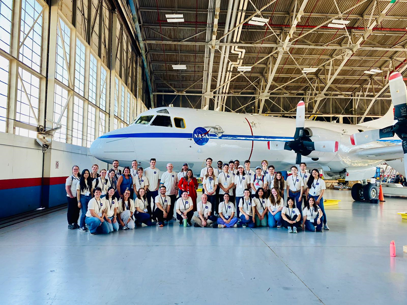 A large white aircraft sits in a hangar with a group of NASA interns posing in front of it. 