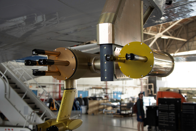 Two scientific instruments which look like copper pipes, sit on the outside of a white wing to an aircraft.