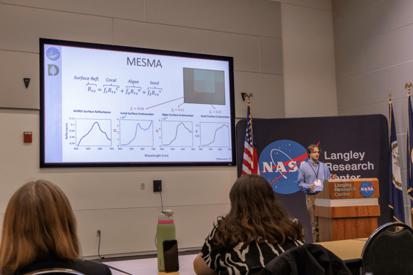 A student gives a presentation behind a lectern while his presentation is projected upon a projection wall. 