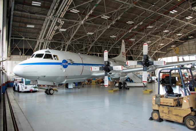 A large white aircraft sits inside a hangar. A forklift sits to the right most edge of the image.