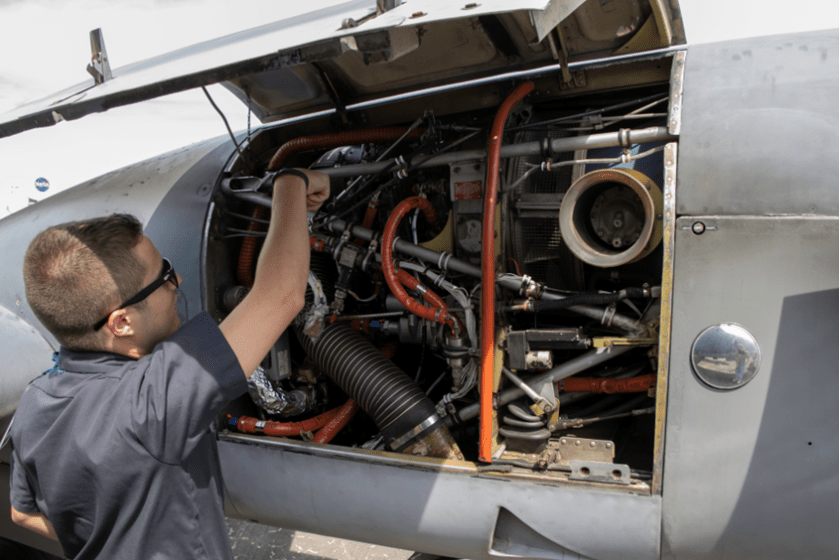 A pilot with sunglasses opens the engine compartment of a gray aircraft.