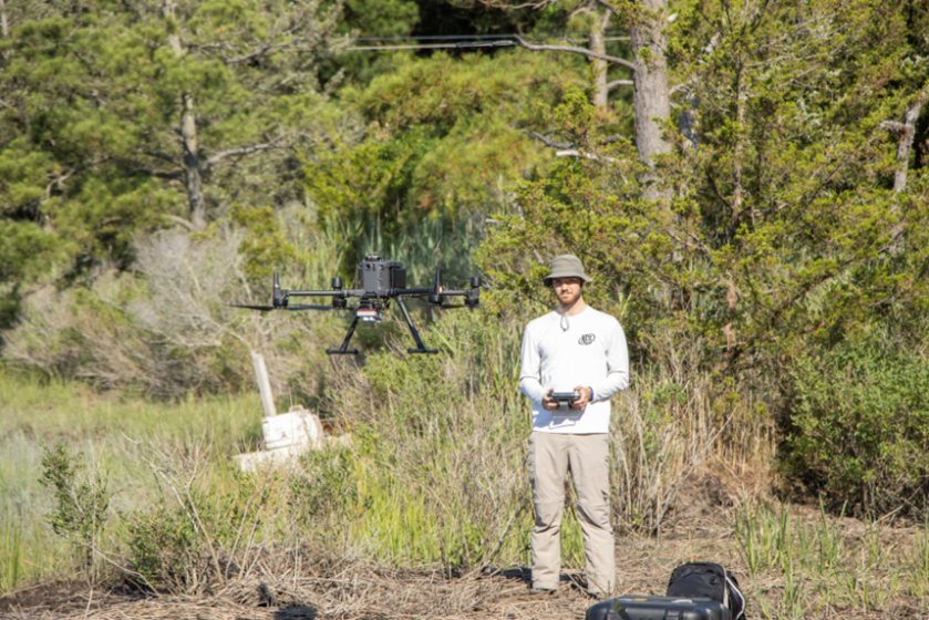 A man operates a quadcopter drone while wearing long sleeve attire and a hat. 