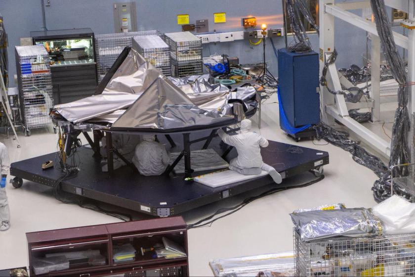 A clean room with assembly workers in white coveralls operating on a large metallic reflective structure. 