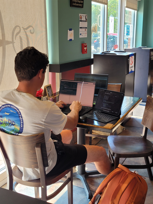 A student sits in a cafe working at a table using multiple laptops. 