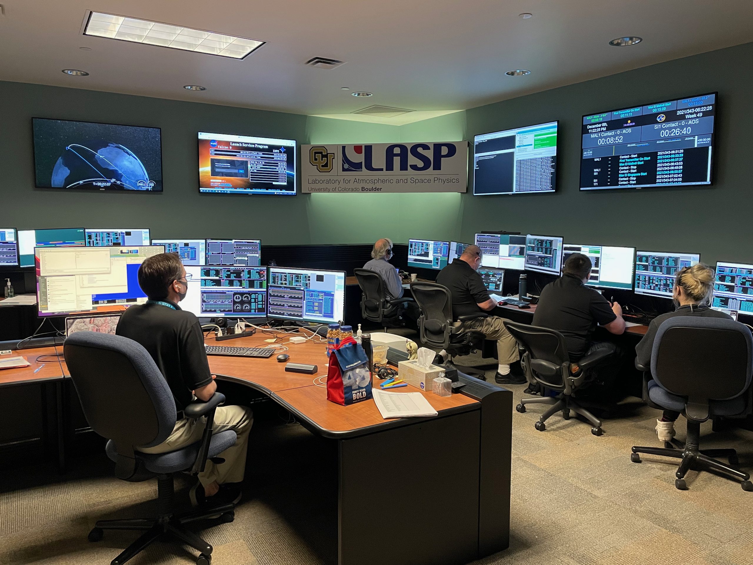 A team of people of LASP sit at computer desks lined against the wall to work on IXPE.