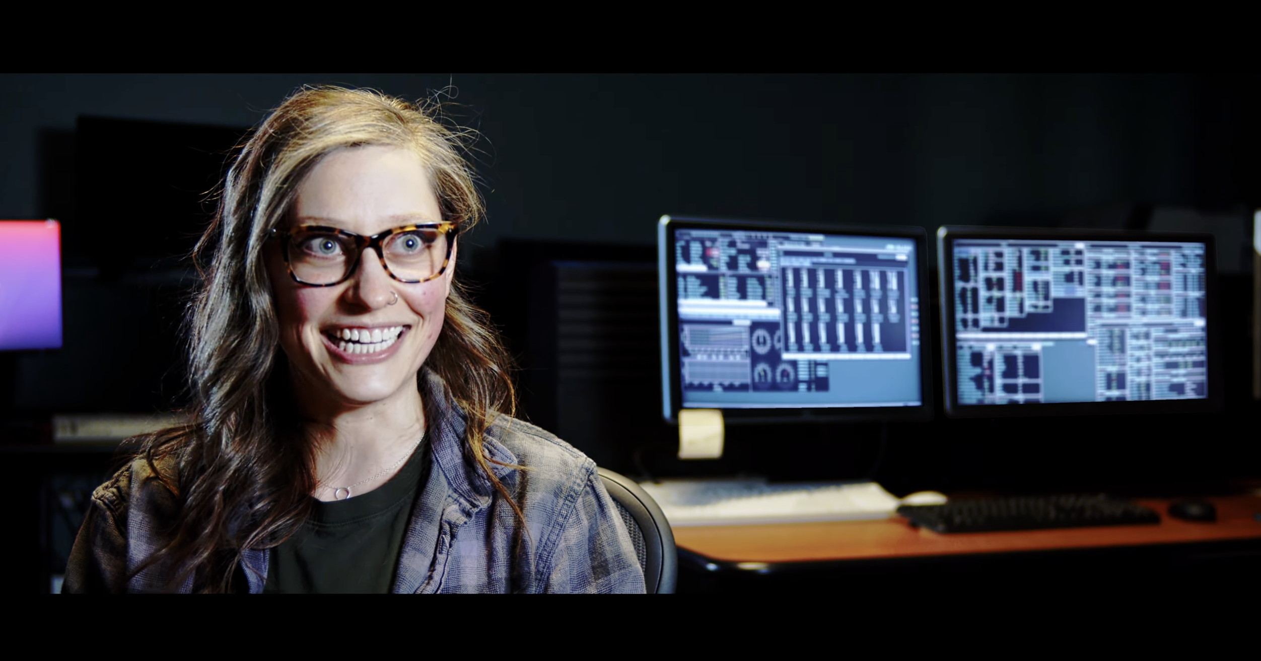 LASP’s Kacie Davis, a women with long hair and glasses, is smiling and is sitting in front of computer screens