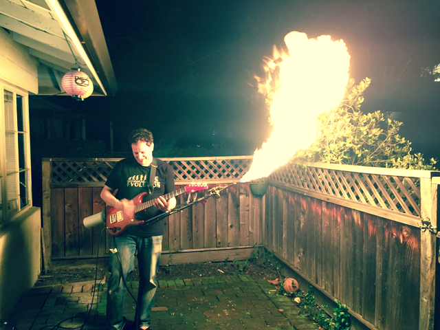 A man plays a flamethrowing guitar in a backyard. 